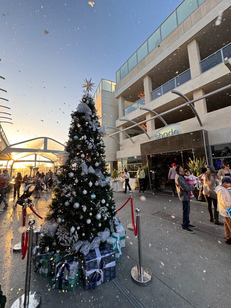 A decorated Christmas tree outside Shade Hotel Redondo Beach during a festive event with families, snow, and gift boxes under the tree at sunset.