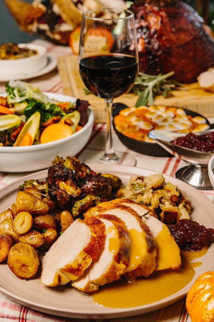 Sea Level Thanksgiving Feast at Shade Hotel Redondo Beach consisting of turkey with gravy, potatoes, brussels sprouts, stuffing, cranberry sauce, sweet potato casserole and a glass of red wine. Honey glazed ham and other side dishes in the background.