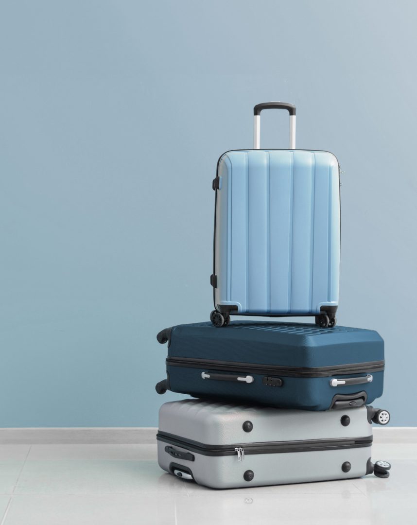 A stack of three modern suitcases in light blue, dark blue, and silver, standing on a white floor against a matching light blue wall background.