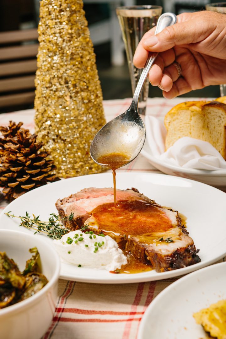 A hand pours gravy over a prime rib dinner served with horseradish and herbs on a holiday table with a gold tree and pinecone decoration for Christmas dinner at Sea Level Restaurant at Shade Hotel Redondo