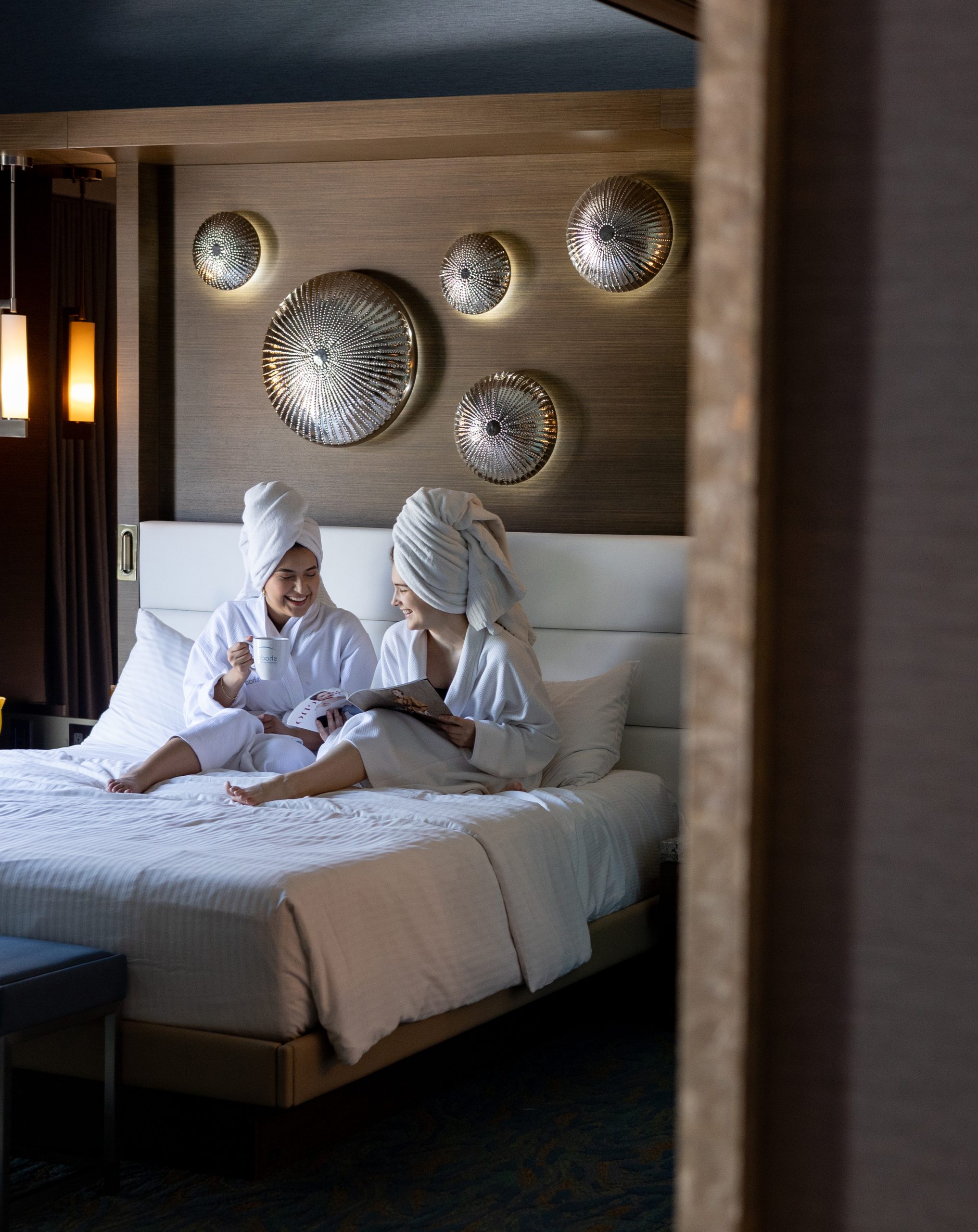 Two women wearing robes and towels on their heads, laughing and drinking coffee in bed at Shade Hotel Redondo Beach